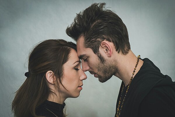 Ein Mann und eine Frau stehen mit geschlossenen Augen eng aneinander, die Stirnen berühren sich. Beide wirken ruhig und nachdenklich, als würden sie einen stillen, emotionalen Moment teilen.