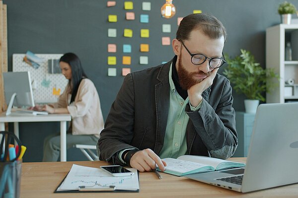 Mann sitzt gelangweilt vor Laptop im Büro, während eine Kollegin im Hintergrund arbeitet. Symbolbild für Langeweile und Unterforderung am Arbeitsplatz.