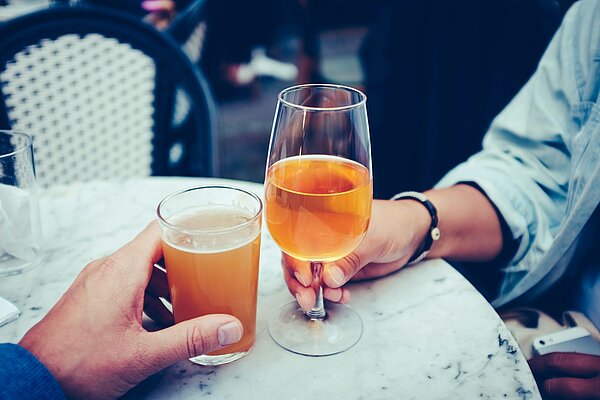 Zwei Menschen stoßen an einem Tisch mit Bier und Wein an – Symbolbild für alltäglichen Alkoholkonsum im sozialen Kontext.