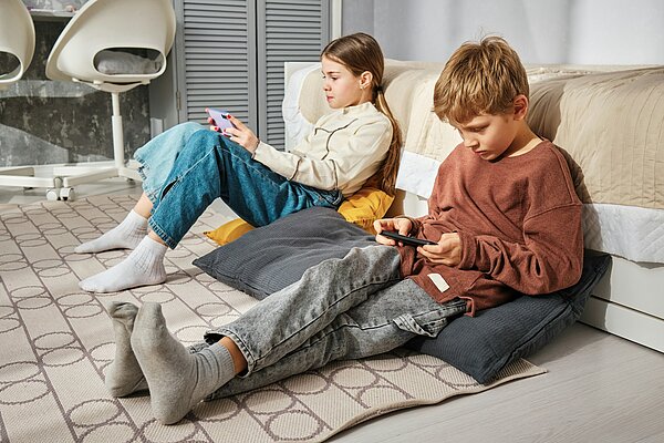 Zwei Kinder sitzen mit Smartphones in der Hand auf dem Boden eines Zimmers und sind konzentriert auf ihre Bildschirme fokussiert.