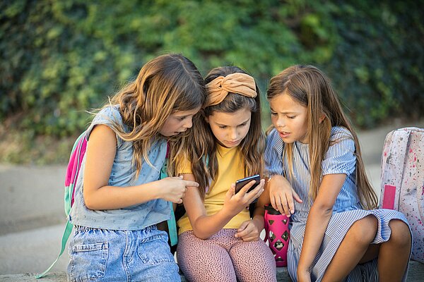 Drei Grundschulkinder sitzen nebeneinander und schauen gemeinsam auf ein Smartphone. Sie wirken neugierig und konzentriert.