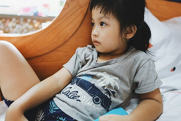 Ein kleines Mädchen liegt nachdenklich auf einem Bett und schaut ausdruckslos zur Seite. Sie trägt ein graues T-Shirt und wirkt still und in sich gekehrt – ein Moment der Langeweile oder inneren Ruhe.