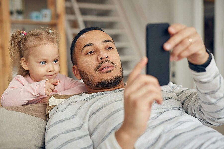 Ein Vater liegt auf dem Sofa und schaut auf sein Smartphone, während seine kleine Tochter neben ihm sitzt und ihn aufmerksam beobachtet. Das Bild zeigt symbolisch, wie Kinder das Verhalten ihrer Eltern wahrnehmen und nachahmen.