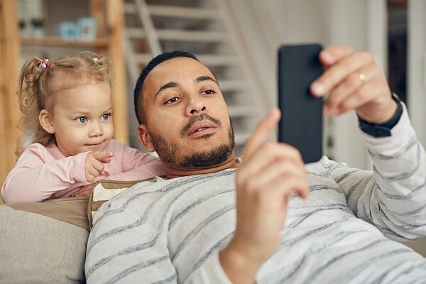 Ein Vater liegt auf dem Sofa und schaut auf sein Smartphone, während seine kleine Tochter neben ihm sitzt und ihn aufmerksam beobachtet. Das Bild zeigt symbolisch, wie Kinder das Verhalten ihrer Eltern wahrnehmen und nachahmen.