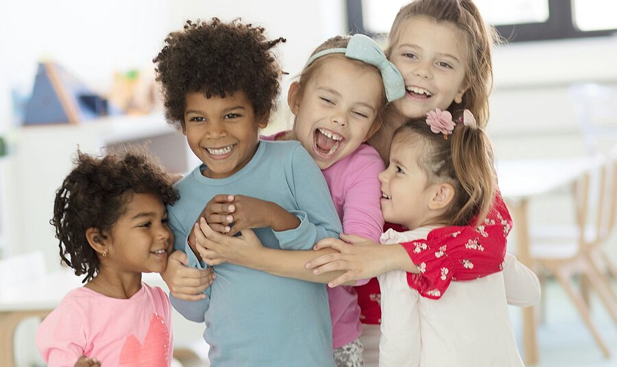 Mehrere fröhliche Kinder umarmen sich lachend in einem hellen Raum – Symbol für Vertrauen, Gemeinschaft und emotionale Sicherheit im Alltag.